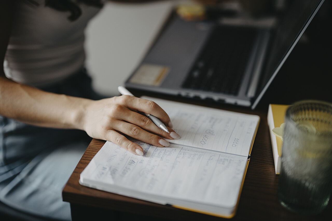 Hands writing in a journal next to a laptop