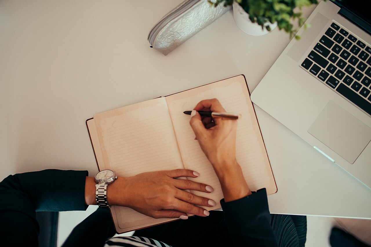 Hands writing in a notebook next to a laptop