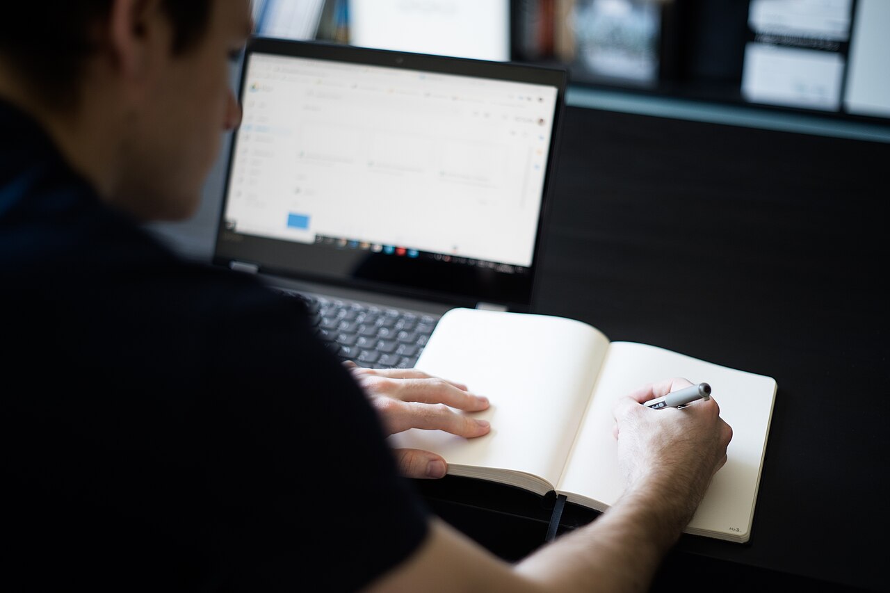 A man writing in a notebook while working on a laptop
